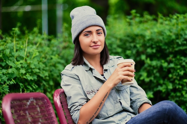 Portrait of brunette female in a hat drinks coffee in a summer park.