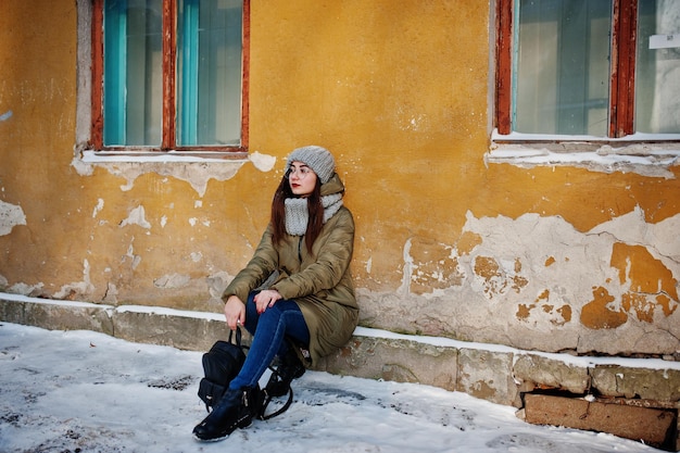 Portrait of brunette girl in gray scarf and hat, glasses at cold weather against orange wall of old house.