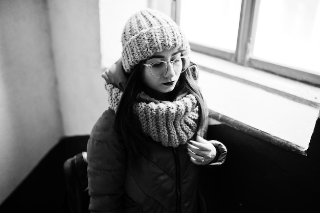 Portrait of brunette girl in gray scarf and hat glasses near window