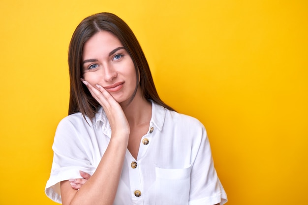 Portrait of brunette girl holding her cheek with hand, is sad and thinks isolated on yellow