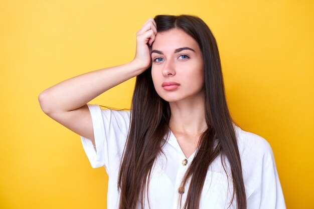 Portrait of brunette girl holding her head, thinking and looking at the camera isolated on yellow background