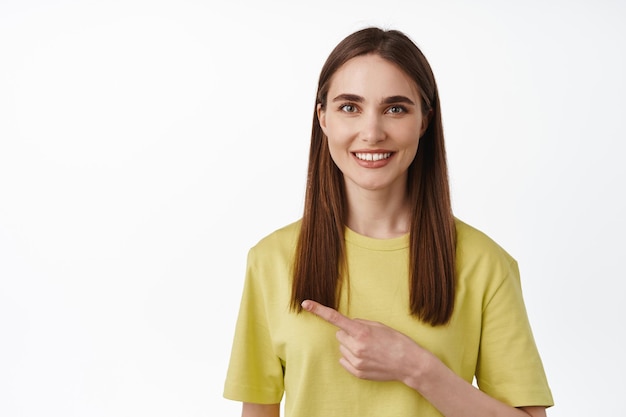 Portrait of brunette girl smiling, pointing left at logo, looking friendly, showing advertisement, recommend something, standing in yellow t-shirt over white background