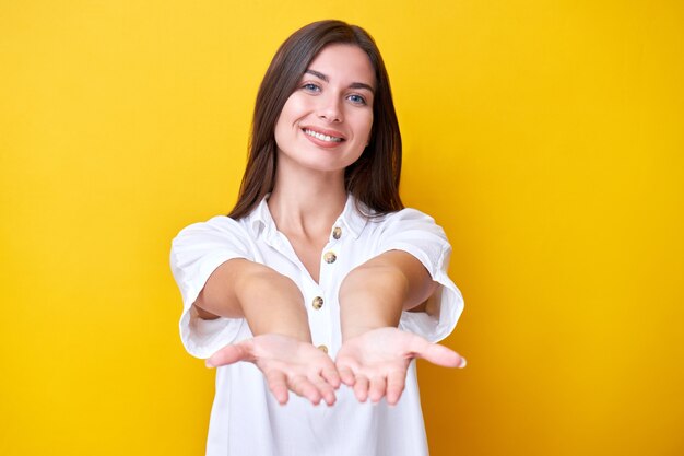 Portrait of brunette girl smiling and stretching hands to the camera with copy space, pretending to hold gift isolated on yellow wall