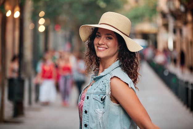 バルセロナの街の通りで写真撮影のためにポーズをとって帽子とバッグを持つブルネットの少女の肖像画。