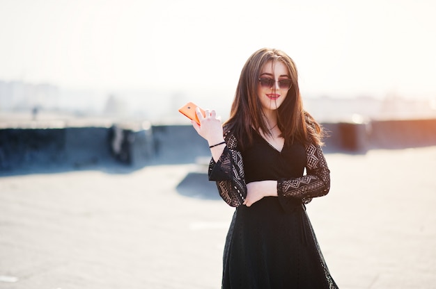 Portrait brunette girl with red lips and orange mobile phone at hands, wearing a black dress, sunglasses posed on the roof