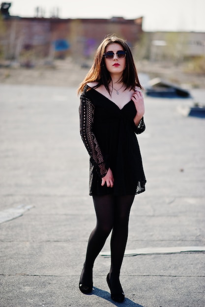 Portrait brunette girl with red lips wearing a black dress, tights and shoes on heels posed on the roof