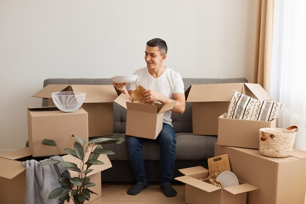 Portrait of brunette handsome man wearing T-shirt and jeans sitting on sofa surrounded with pile of packages, unpack personal stuff from carton boxes, being happy plate is not broken during relocating