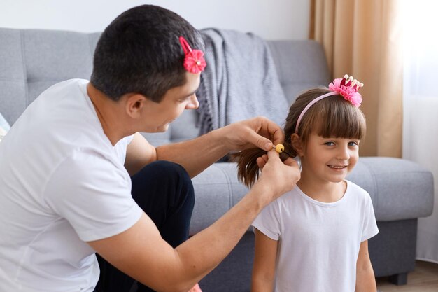 Portrait of brunette man spending time with his daughter, making pigtails while sitting on floor near sofa, smiling cute girl with charming smile looking at camera.