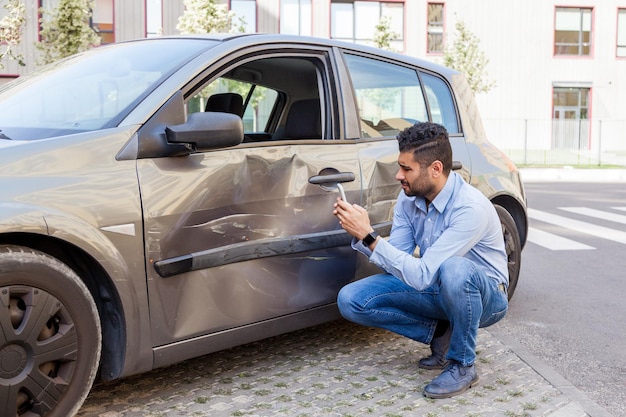 Photo portrait of brunette man wearing jeans and blue shirt not cope with driving damaged his car door dents and scratches on auto body making photos of damaging for insurance inspection outdoor shot