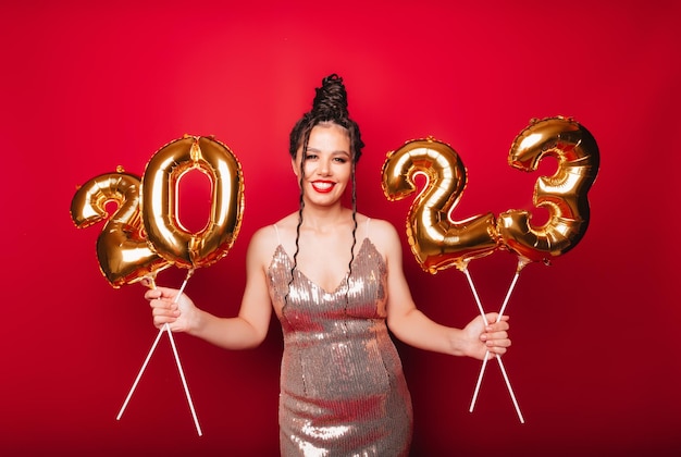 Portrait of a brunette on a red background holding golden balloons in the shape of 2023 Coming New Year Christmas space for text