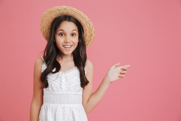 Portrait of brunette smiling girl 8-10 wearing white dress and straw hat looking at camera and pointing finger aside at copyspace, isolated over pink background