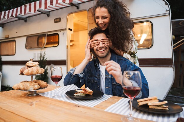 Portrait of brunette smiling woman with curly long hair covering her boyfriend‘s eyes at dinner near trailer outdoors