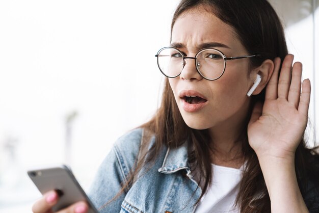 Portrait of brunette teenage girl wearing earpods and eyeglasses holding her hand at ear and trying to listen while using smartphone outdoors