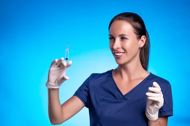 Portrait of a brunette with a braided tail, posing while standing against a blue background, in a blue surgical suit, in medical gloves, squeezing a medicine from a syringe.