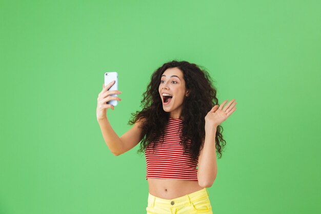 Portrait of brunette woman 20s wearing summer clothes smiling and using cell phone isolated on green