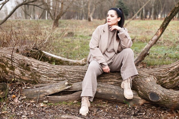 Portrait of brunette woman in beige coat with beautiful long hair sitting on a tree in the park