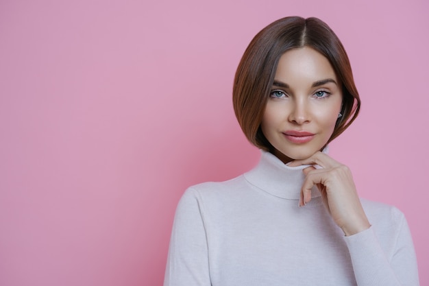 Portrait of brunette woman keeps hand on collar of turtleneck, has healthy perfect skin