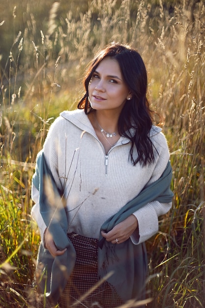 Portrait of a brunette woman in a sweater and scarf in autumn in a field with dry grass at sunset