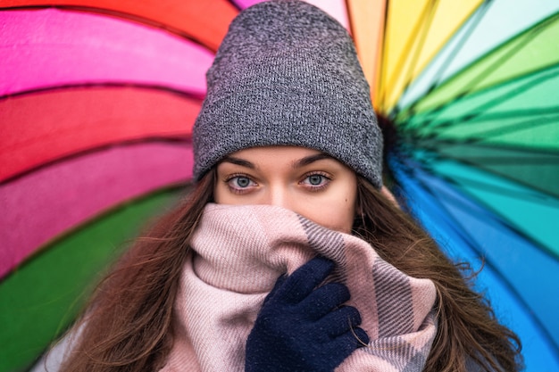 Ritratto di donna bruna con bellissimi occhi felici avvolti in sciarpa calda e abiti accoglienti con ombrello arcobaleno multicolore luminoso