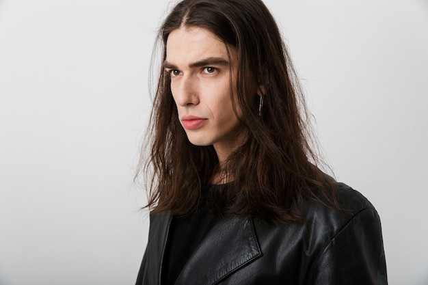 Portrait of brunette young man with long hair wearing black lather jacket posing and looking aside on white