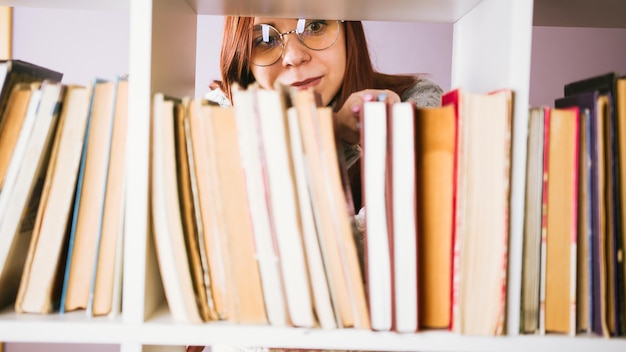 Портрет брюнетки, пытающейся найти книги для чтения Женщина в очках ищет книгу на книжных полках в библиотеке Студент смотрит на книжные полки, чтобы найти книгу для чтения