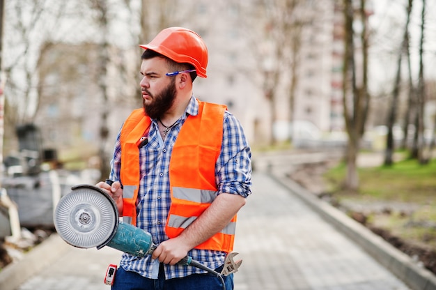 Portrait of brutal beard worker man suit construction worker in safety orange helmet against pavement with angular grinding machine in hand.