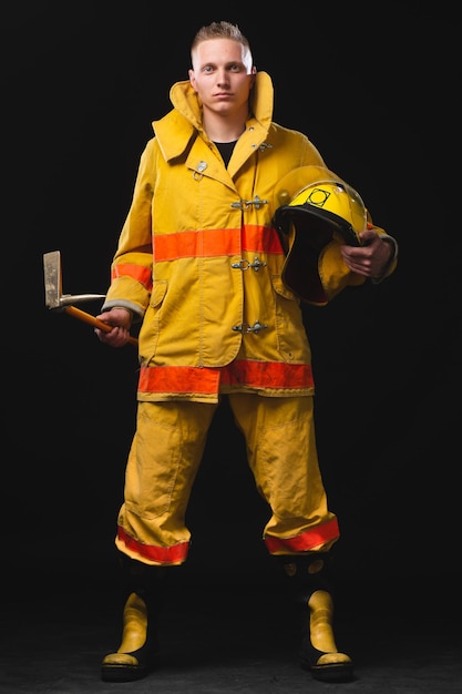 Foto ritratto di vigile del fuoco brutale che tiene casco e giacca in piedi in studio contro il muro scuro