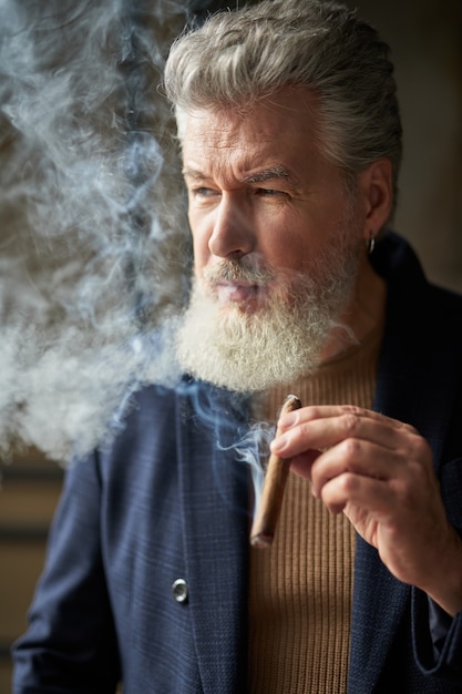Портрет брутального седого зрелого мужчины, смотрящего в сторону с сигарой в руке