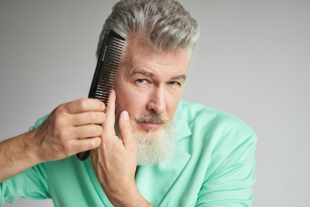 Portrait of brutal middle aged man with beard looking at camera brushing his hair with