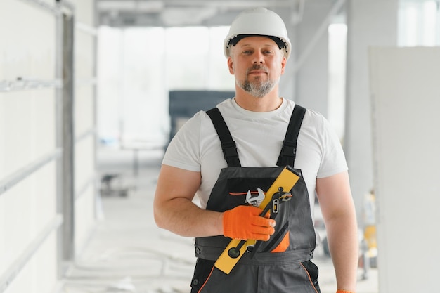 屋内の建設現場で器具を持って立つ、保護ヘルメットをかぶった作業服を着たビルダーのポートレート