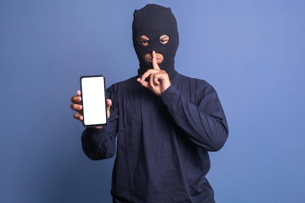 Portrait of a burglar putting a finger on his lips asking for silence while showing smartphone with