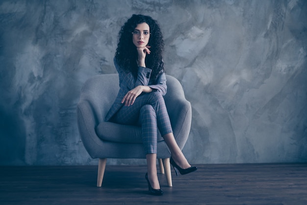 Portrait of business lady sitting in chair with crossed legs isolated over concrete wall