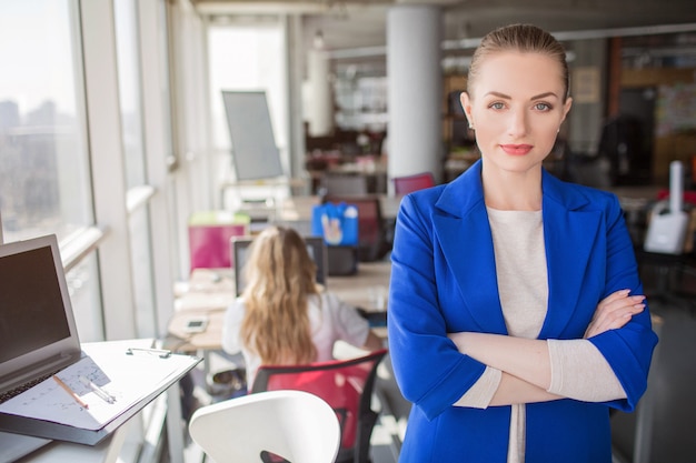 A portrait of business lady standing near the window with her hands crossed. She is looking straight to the camera. Girl looks confident and very good.