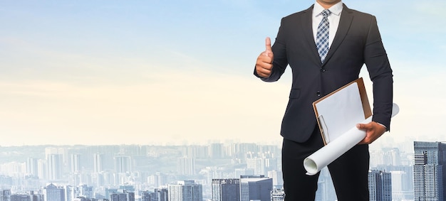 Portrait of business man wearing a black suit holding a blueprint isolated on city background