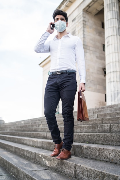 Portrait of business man wearing face mask and talking on the phone while standing on stairs outdoors. Business concept. New normal lifestyle concept.