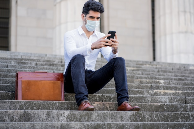 Portrait of business man wearing face mask and using his mobile phone while sitting on stairs outdoors at the street