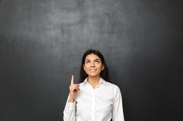 Portrait of business young woman wearing white shirt pointing finger upward on copy space, isolated over dark gray wall