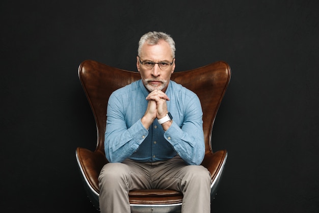 Portrait of businesslike gentleman 50s with grey hair and beard in glasses sitting on wooden armchair with severe look, isolated over black wall