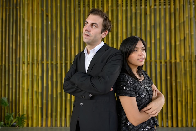 Портрет бизнесмена и азиатской бизнес-леди как многоэтническая пара вместе против бамбуковой стены на открытом воздухе