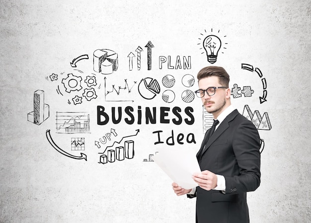 Portrait of a businessman in glasses holding a document and standing near a concrete wall with a black business idea sketch on it.