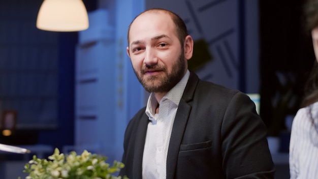 Portrait of businessman looking into camera sitting at conference table working in meeting room late at night. Diverse multi ethnic teamwork analyzing company marketing strategy brainstorming ideas