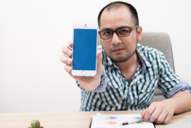 Portrait of businessman sitting behind table showing smartphone with blue screen isolated on white background.