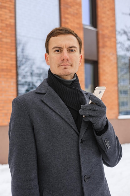 Портрет бизнесмена со смартфоном, стоящего зимой на городской улице и смотрящего в камеру