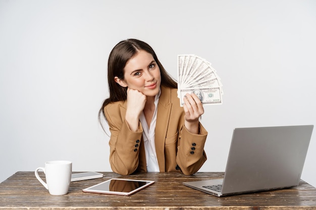 Portrait of businesswoman sitting in office with money, working and making profit income, posing happy against white studio background.