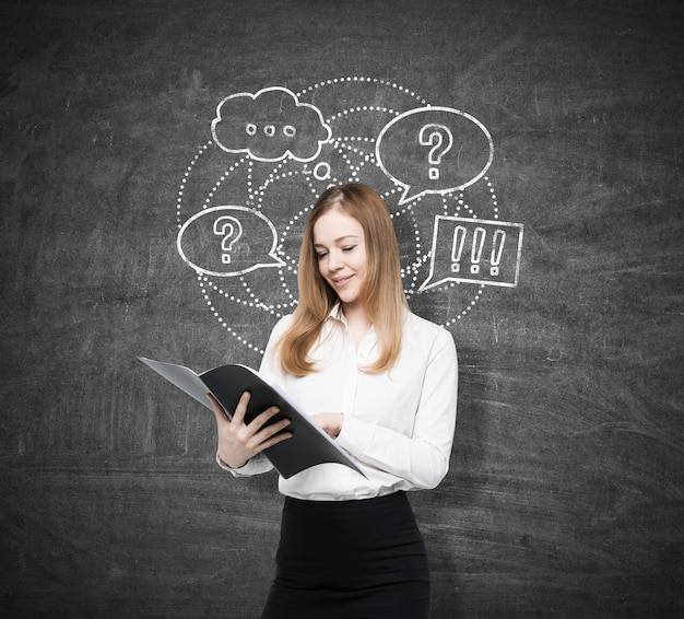 Portrait of a businesswoman with a folder. She is reading a document while standing near a blackboard with a round question and answer sketch