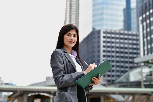 Portrait of businesswoman writing on clipboard while standing against buildings in city