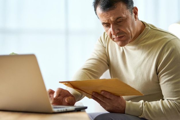 Portrait of busy latin middle aged businessman looking at the envelope using laptop while working