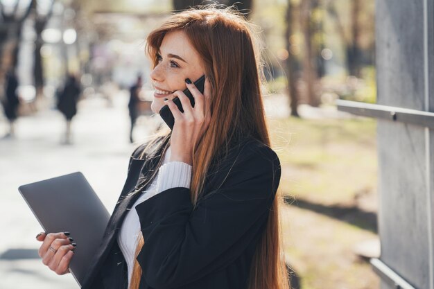 Photo portrait of busy woman freelancer with laptop calling on mobile phone while looking away in park business success business portrait beauty portrait sunny day