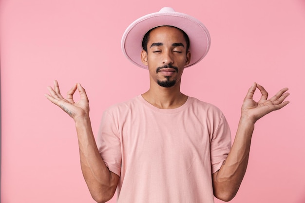 Portrait of calm african american man wearing hat meditating and holding fingers together isolated on pink