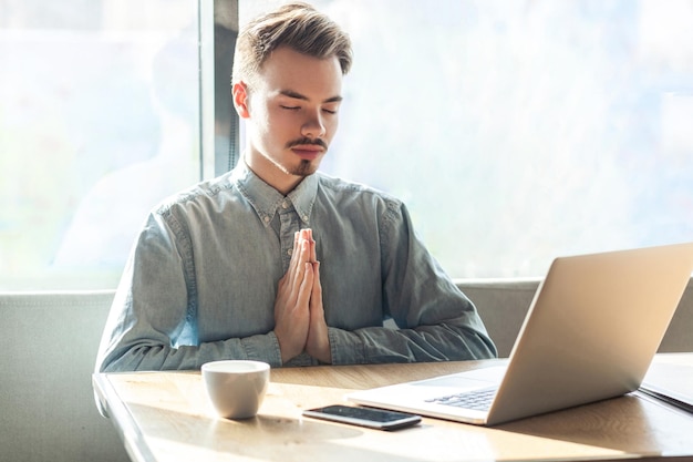 Portrait of calm handsome successful bearded young freelancer in blue shirt are sitting in cafe and having a rest, holding hands like have meditative to have emotional pleasure. indoor